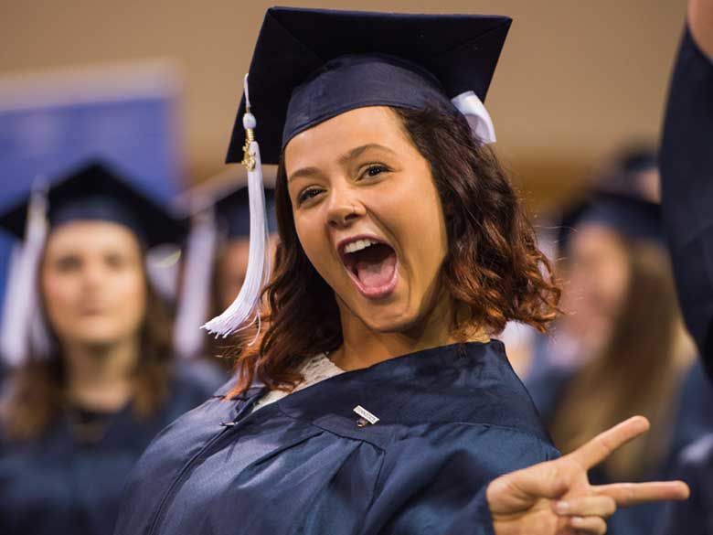 A female graduate in cap and gown makes a victory-sign gesture at commencement.